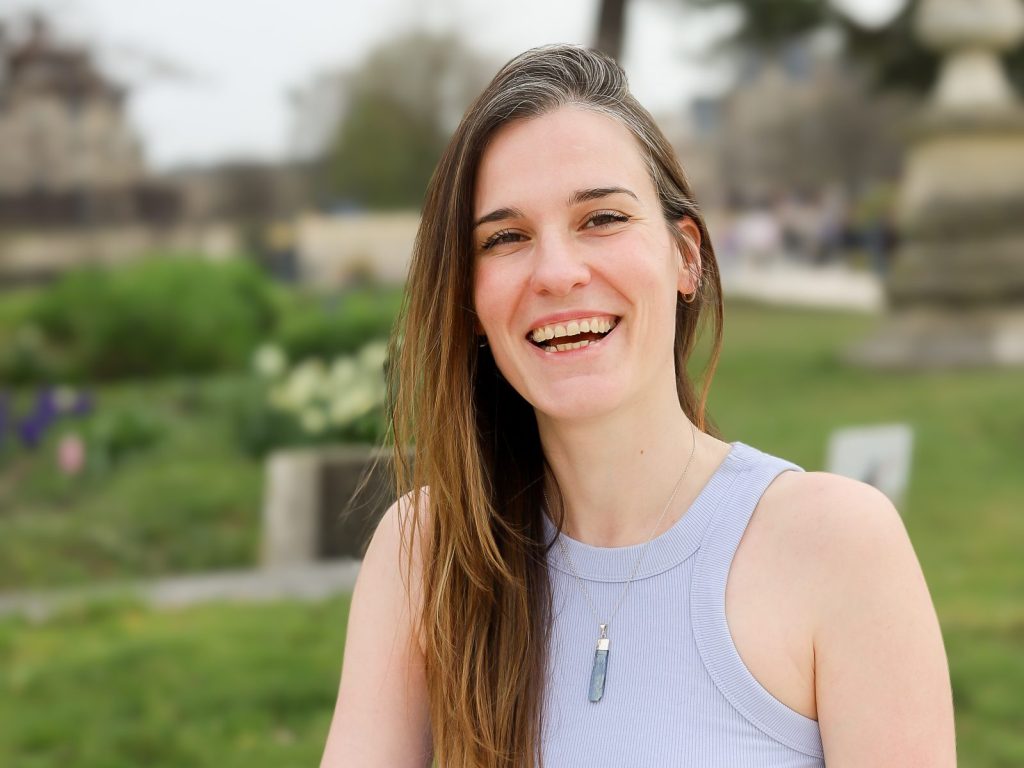 Photo de Vanessa Adis, sourire et regard direct, assise dans un décor arboré et fleuri